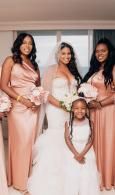 Four people in matching blush dresses pose indoors, one seated in a white gown holding a bouquet.