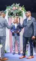 Two men in suits stand under a floral arch with a smiling man holding a book during a ceremony