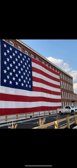 Large American flag displayed on the side of a brick building with a blue sky in the background.