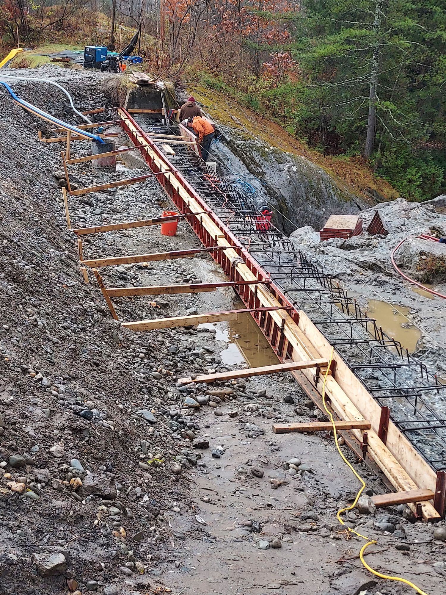 Construction workers building a long concrete channel on a rocky hillside; orange safety vests, brown rock.