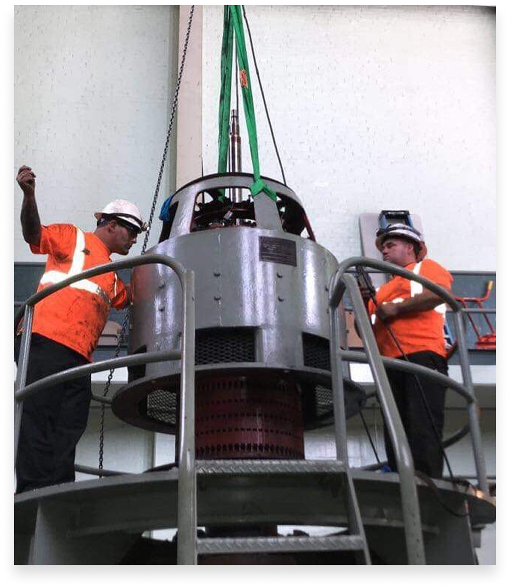 Two workers in orange vests and hard hats inspect a large industrial machine being lifted in a factory.
