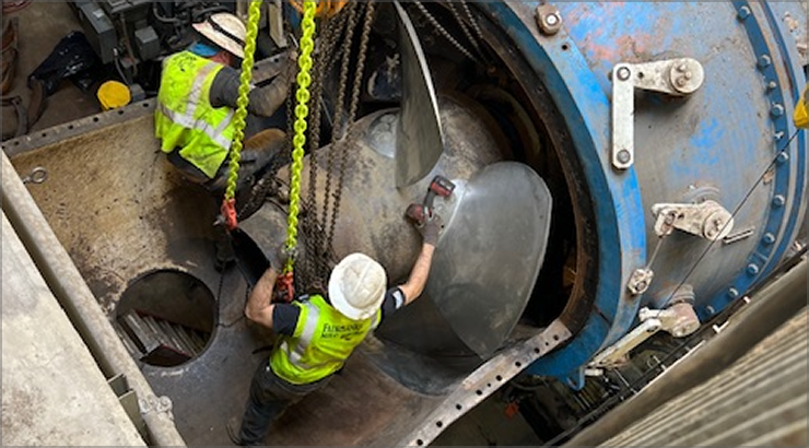 Two workers in safety vests and hard hats inspecting a large blue turbine with metal blades.