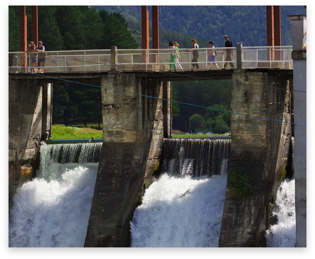 People walking on a bridge over a dam; water gushes through the openings.