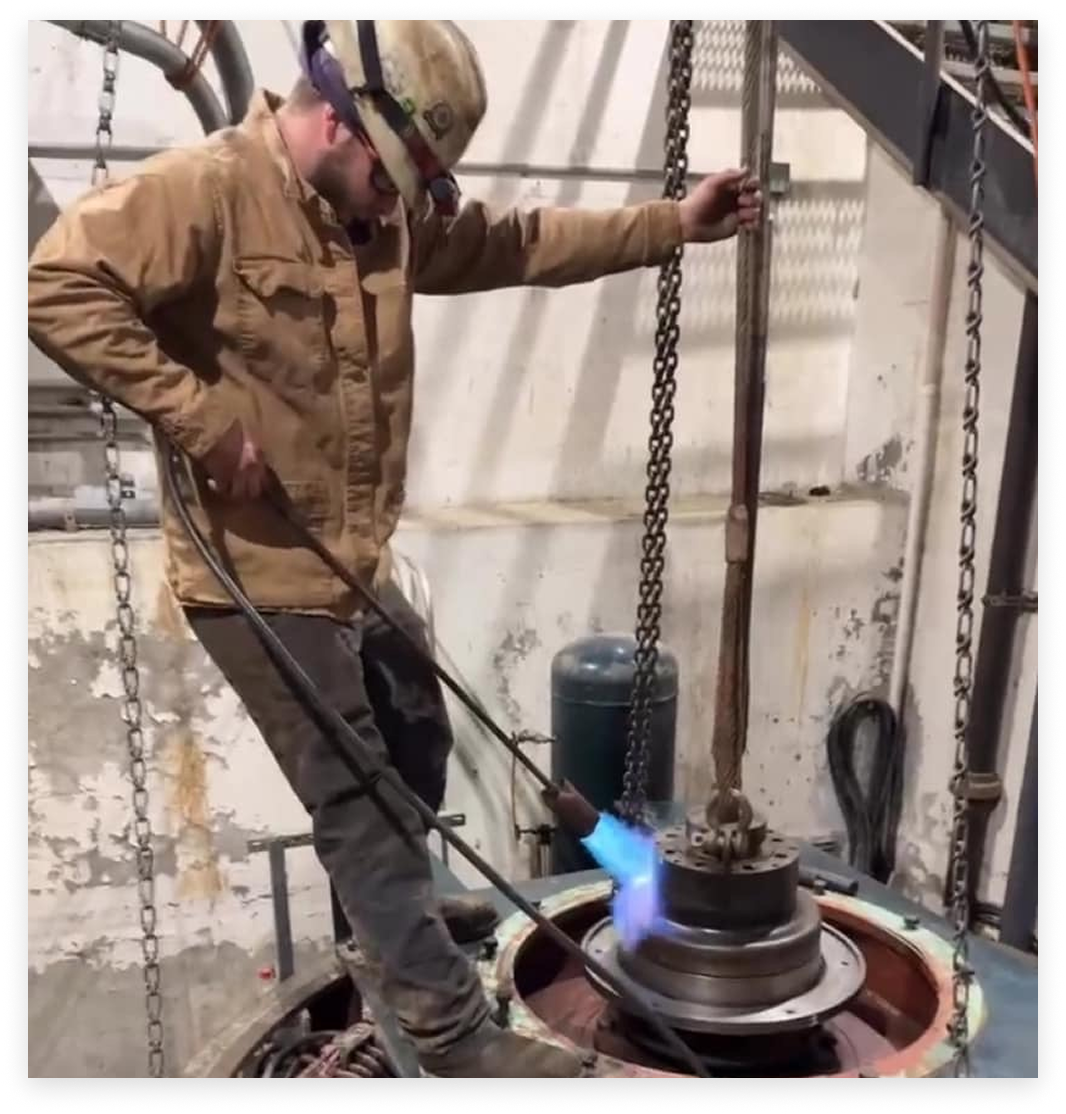 A worker in a welding helmet uses a torch on machinery. Blue flames, chains, and metal elements.