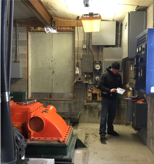 Man in a utility room checks equipment near orange machinery and electrical panels.