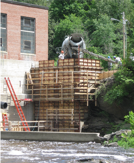 Construction site: concrete being poured into wooden forms near a brick building and river.