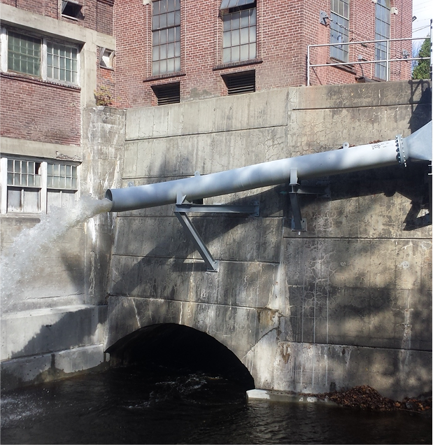 Concrete water passage with a metal pipe; water flowing into a dark tunnel, brick building in background.