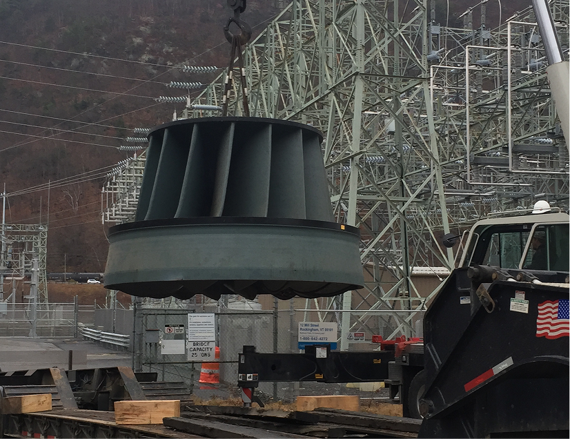Large turbine being lifted by crane at a power plant with transmission towers in the background.