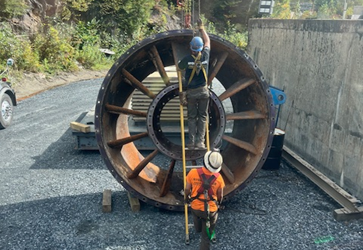 Two workers near a large, rusted turbine wheel, likely for repairs, outdoors.