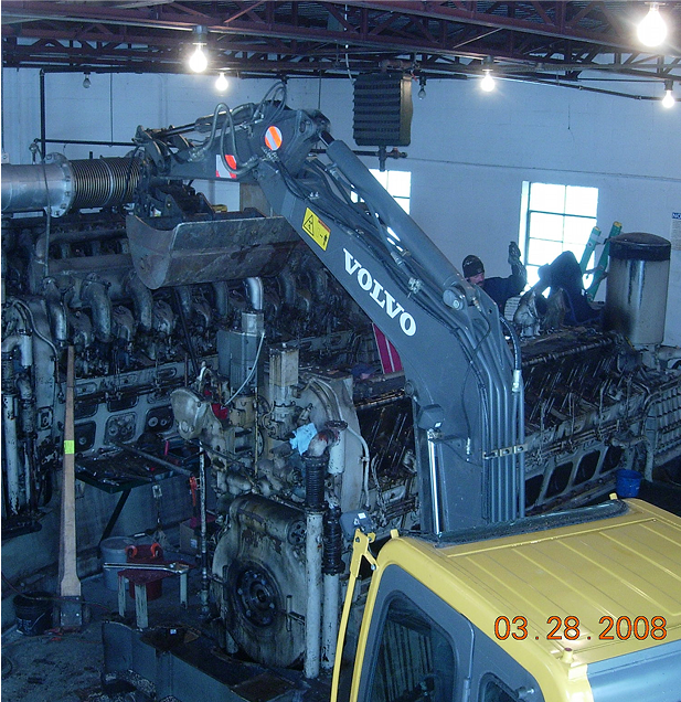 Volvo excavator arm lifting large, complex engine in a shop setting; date stamp reads 