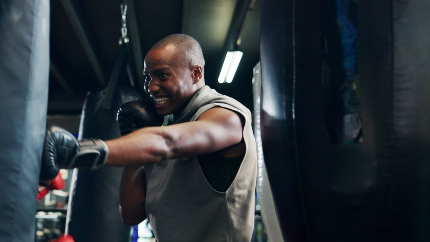 Man boxing a heavy bag in a gym, wearing gloves and smiling while throwing a punch.