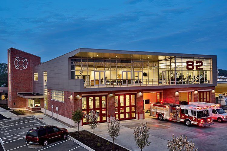 Modern brick and glass fire station numbered 82, with a fire engine parked in front under an evening sky.