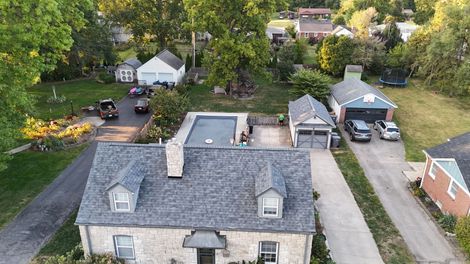 Aerial view of a stone house with a shingled roof, a swimming pool, and garages on a residential property with trees.