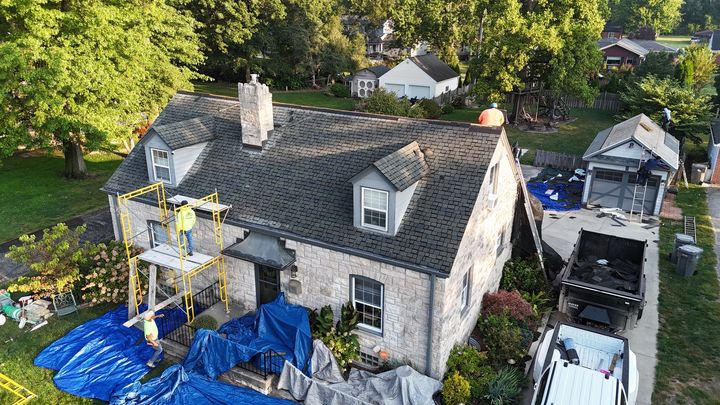 An aerial view shows roofers replacing the asphalt roof of a stone house with scaffolding and blue tarps below.