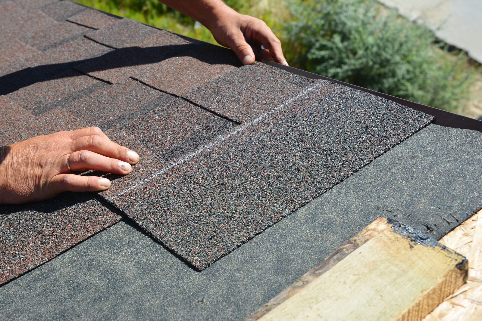 Hands installing asphalt roof shingles on a dark surface, outdoors.