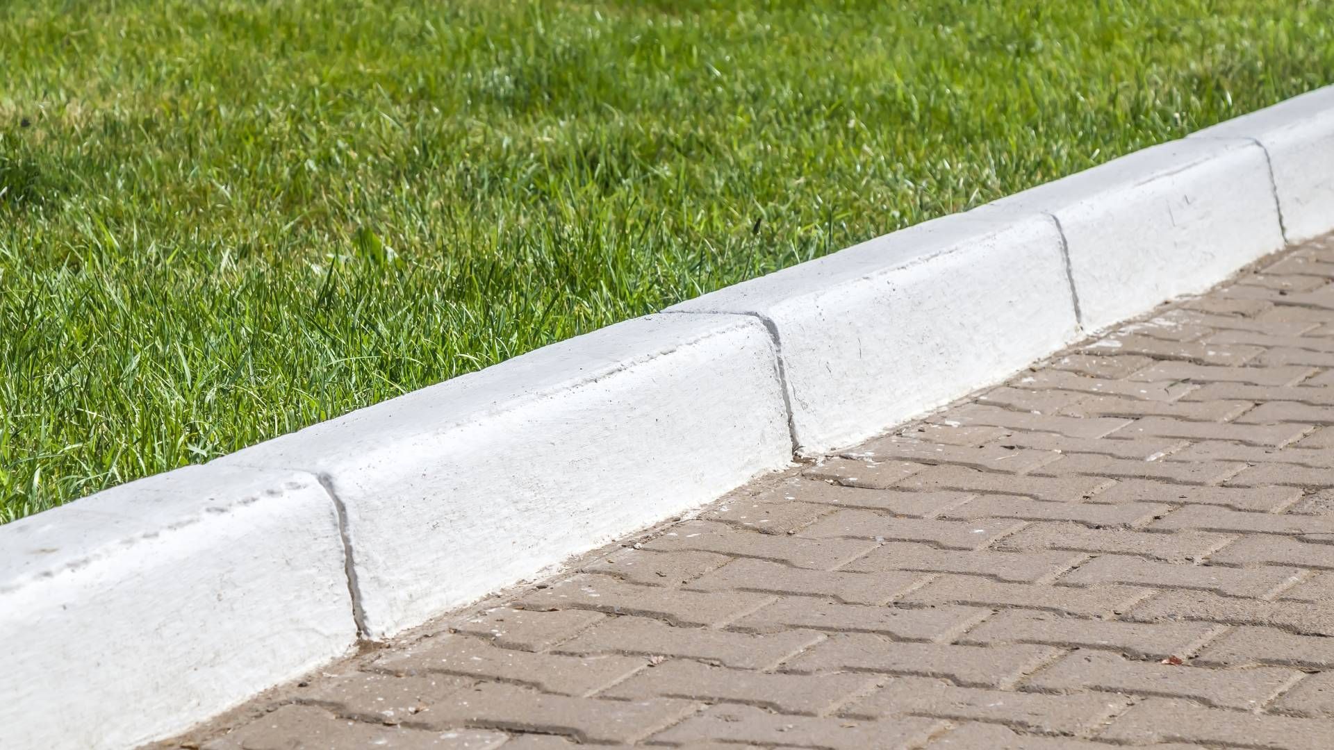 A white curb on a brick sidewalk next to a lush green lawn.