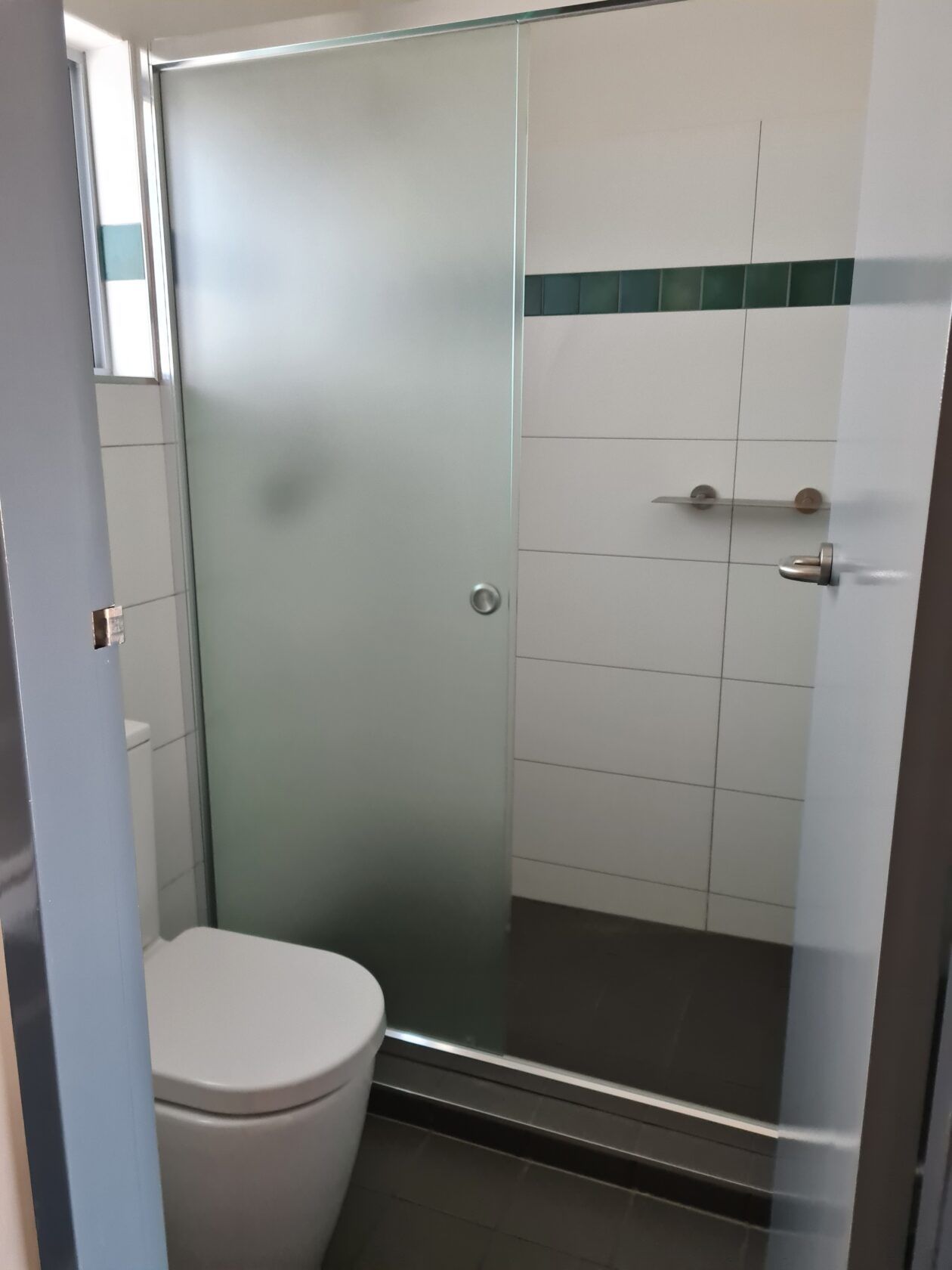 Small bathroom with a frosted glass shower door, toilet, and white tiled walls with a green accent.