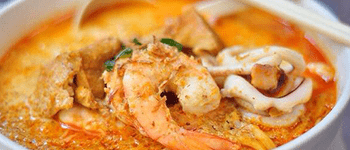 A close-up of a bowl of laksa, a Southeast Asian noodle soup. The broth is an orange color and filled with seafood, tofu and noodles.