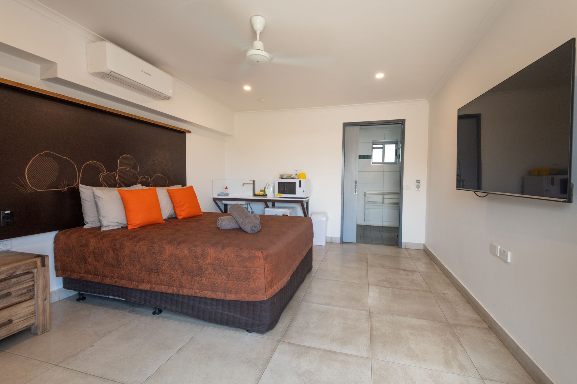 A studio apartment with a bed, TV, kitchenette, and bathroom. The room is white with brown accents, including the bedspread and headboard.