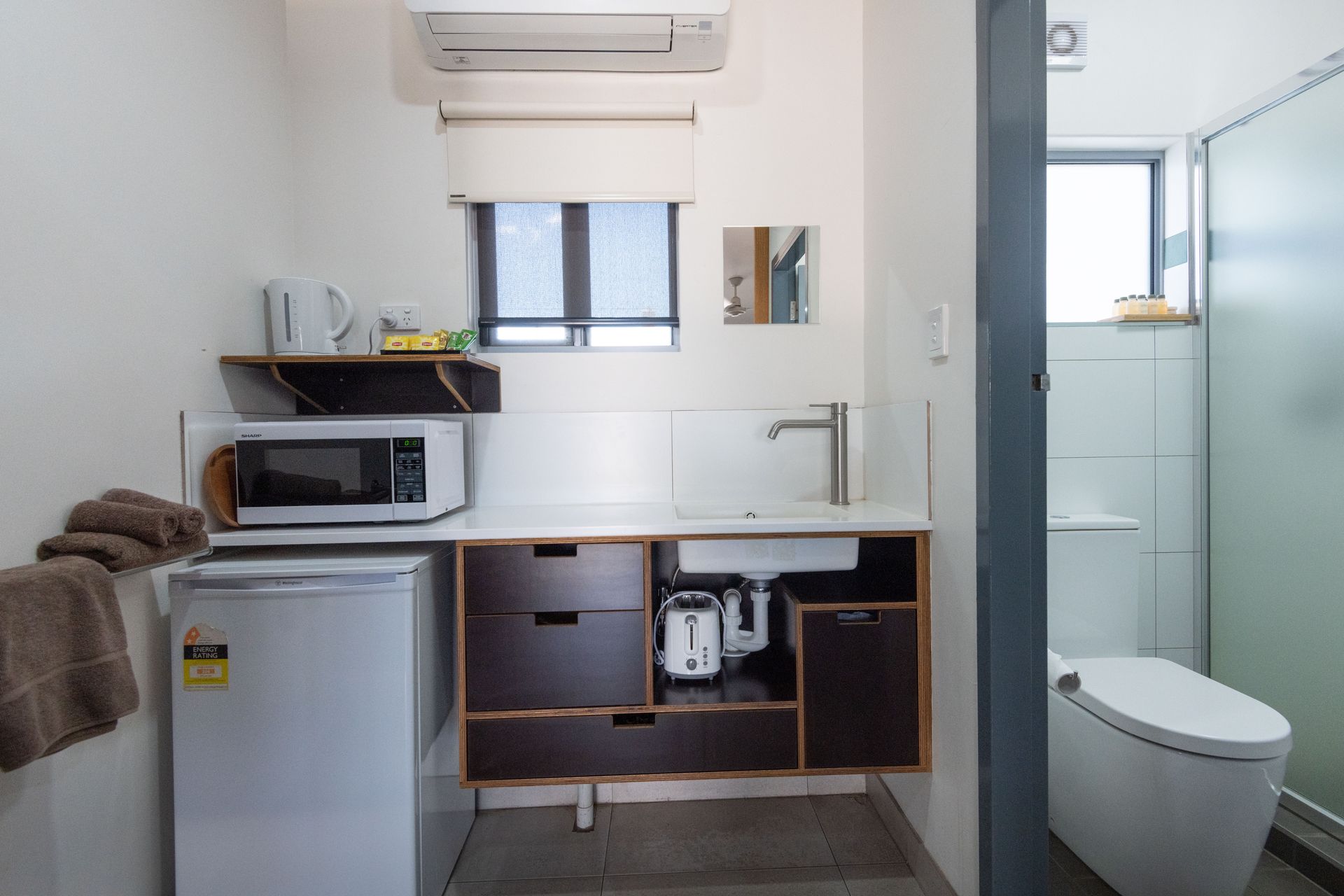 Small kitchenette with a microwave, mini-fridge, sink, and toilet; a small, bright, minimalist space.