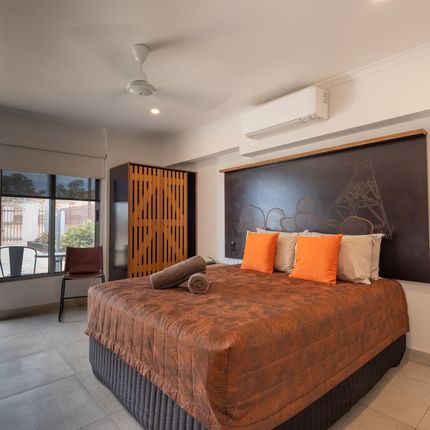 A hotel bedroom with a large bed covered in an orange blanket. There are orange and beige pillows, a window, and a wooden wardrobe.