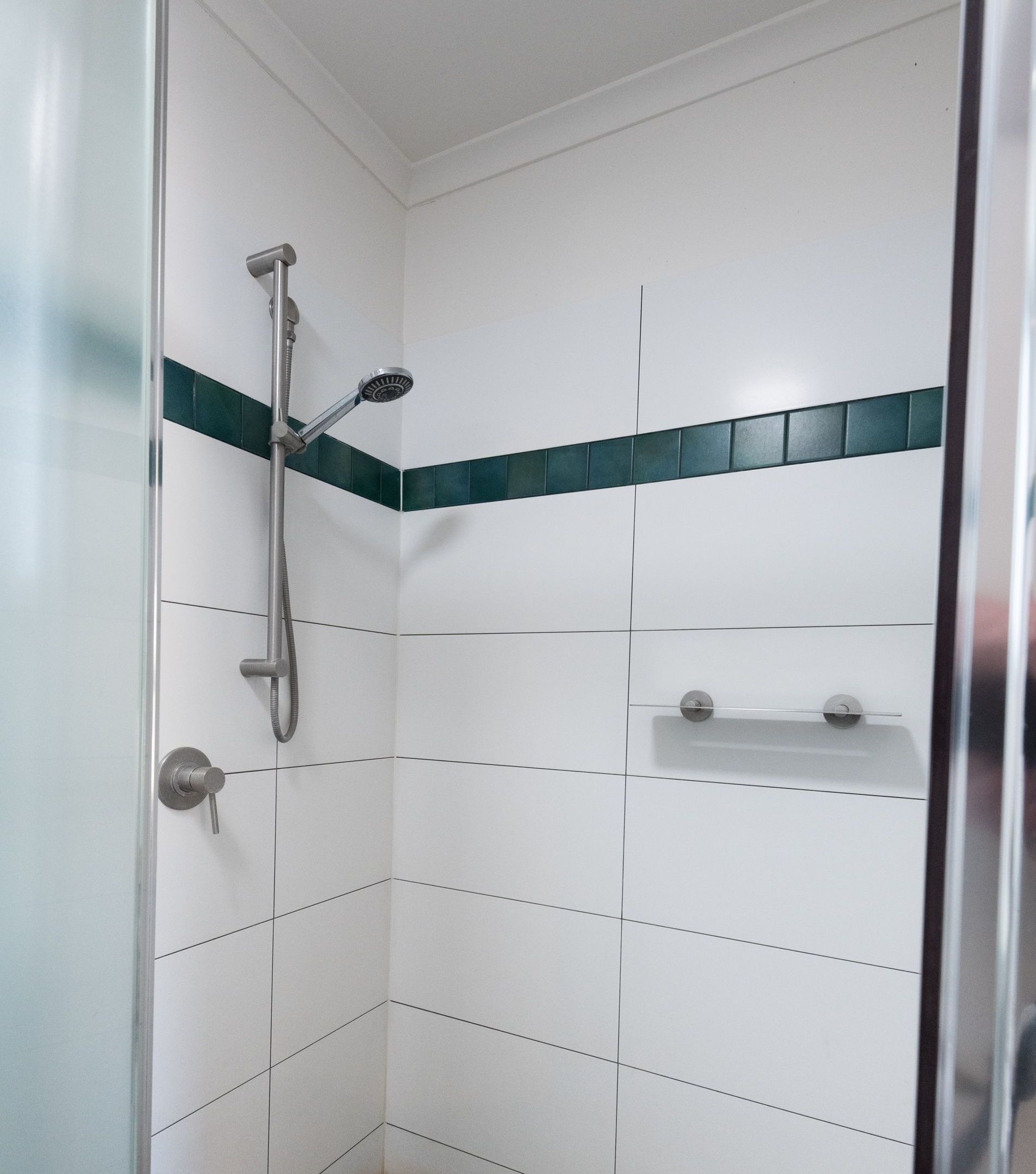 Shower with white walls, a dark green stripe, and a silver shower head. There is a shelf, and the shower is partially enclosed by a glass door.