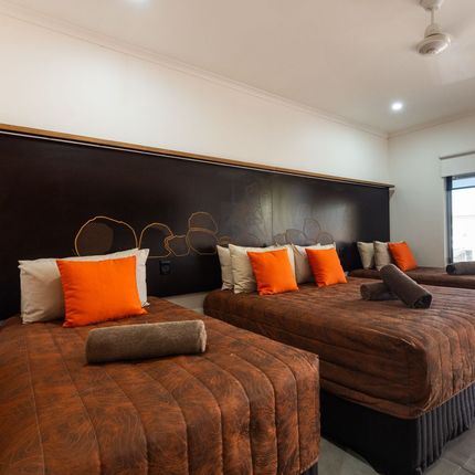 A hotel room with three beds. Brown quilted bedspreads are topped with orange and white pillows.