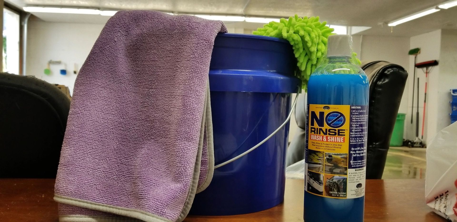 A purple cloth, blue bucket, and blue cleaning solution on a wooden surface. A green wash mitt is attached to the bucket.