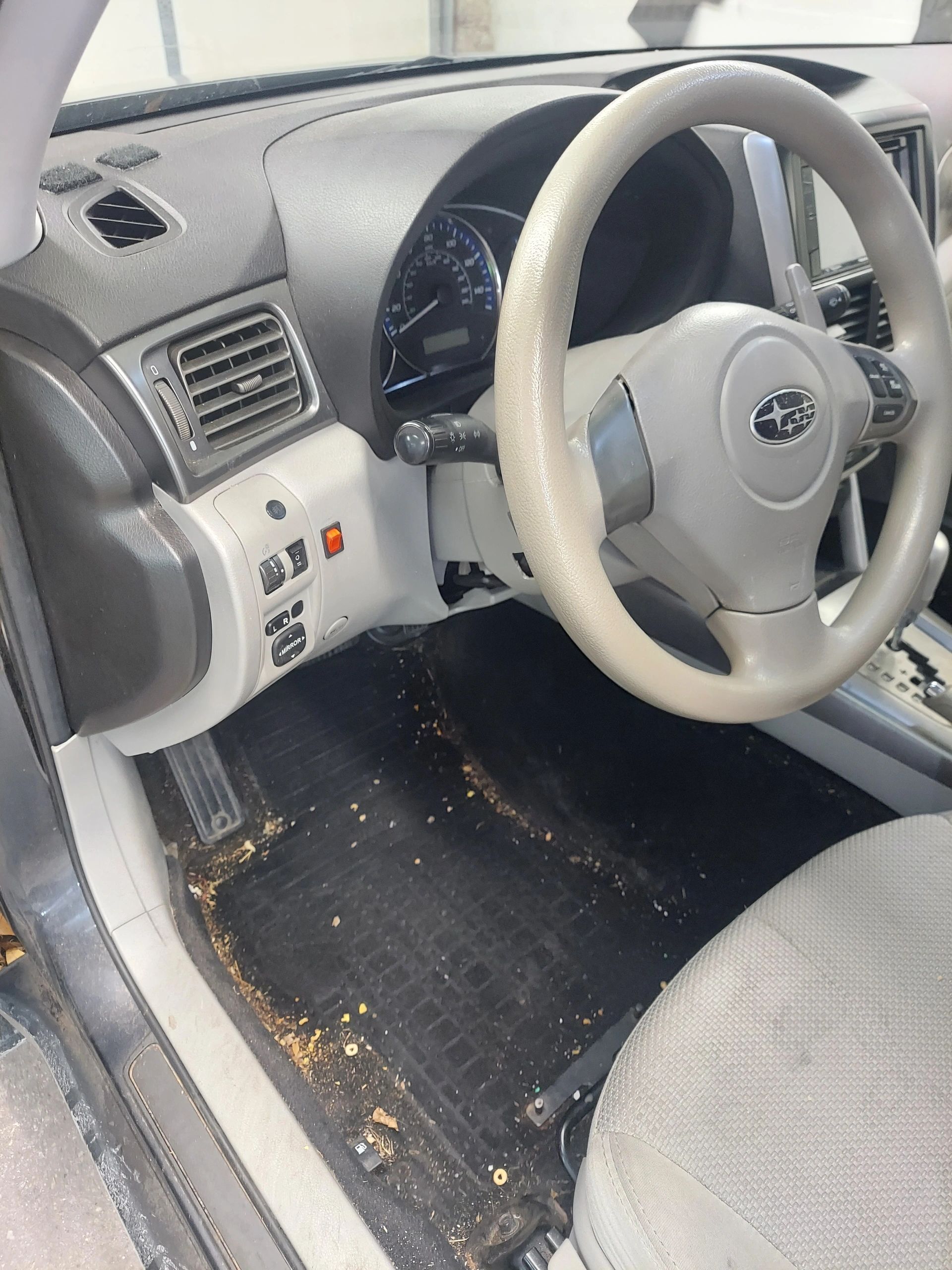 Interior view of a dirty Subaru car. Dusty black floor mats, gray dashboard, beige steering wheel.