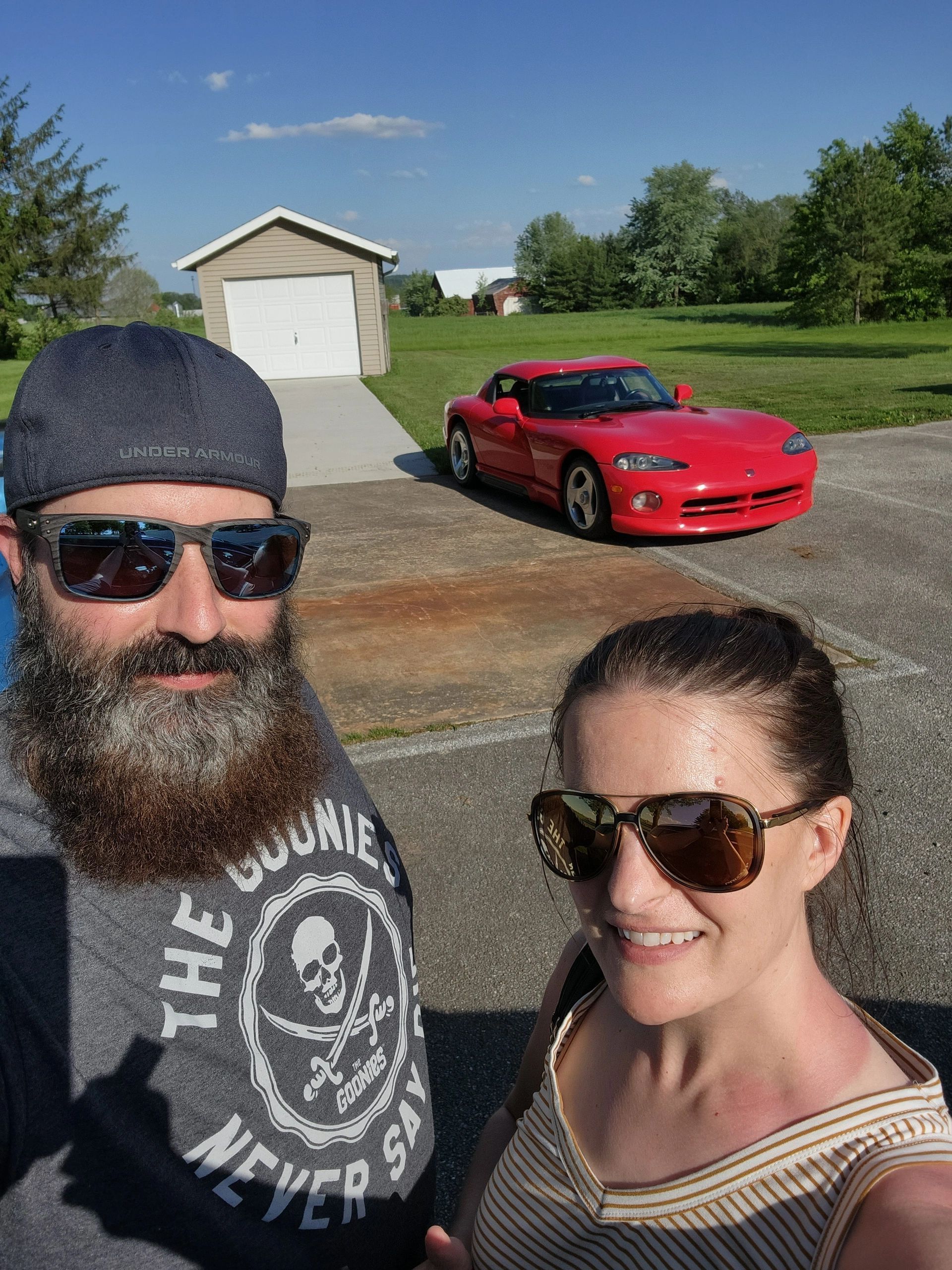 Man with beard and woman smiling in sunglasses, with a red Dodge Viper in front of a garage on a sunny day.