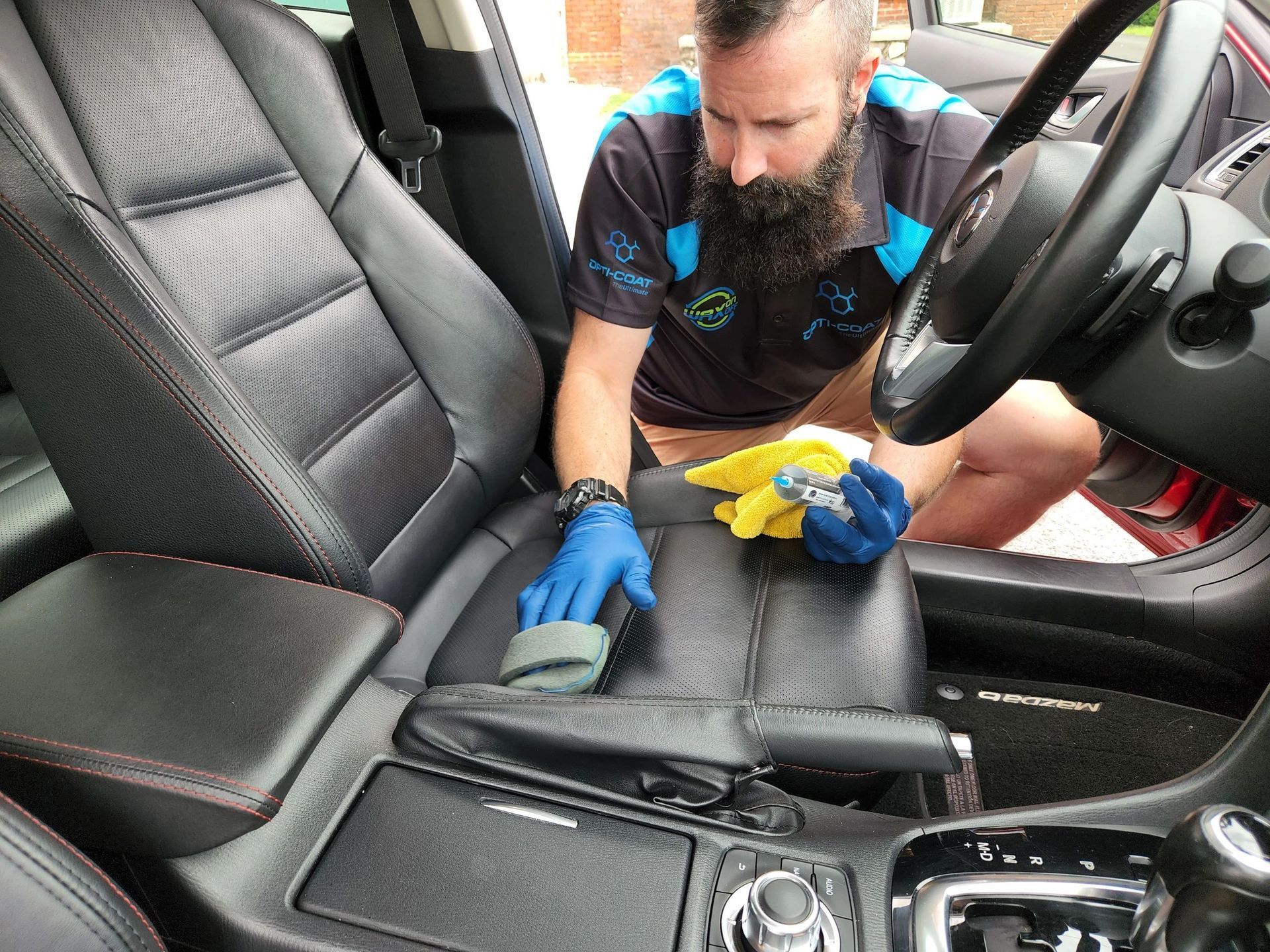 Man cleaning black leather car seat with a sponge, wearing blue gloves.