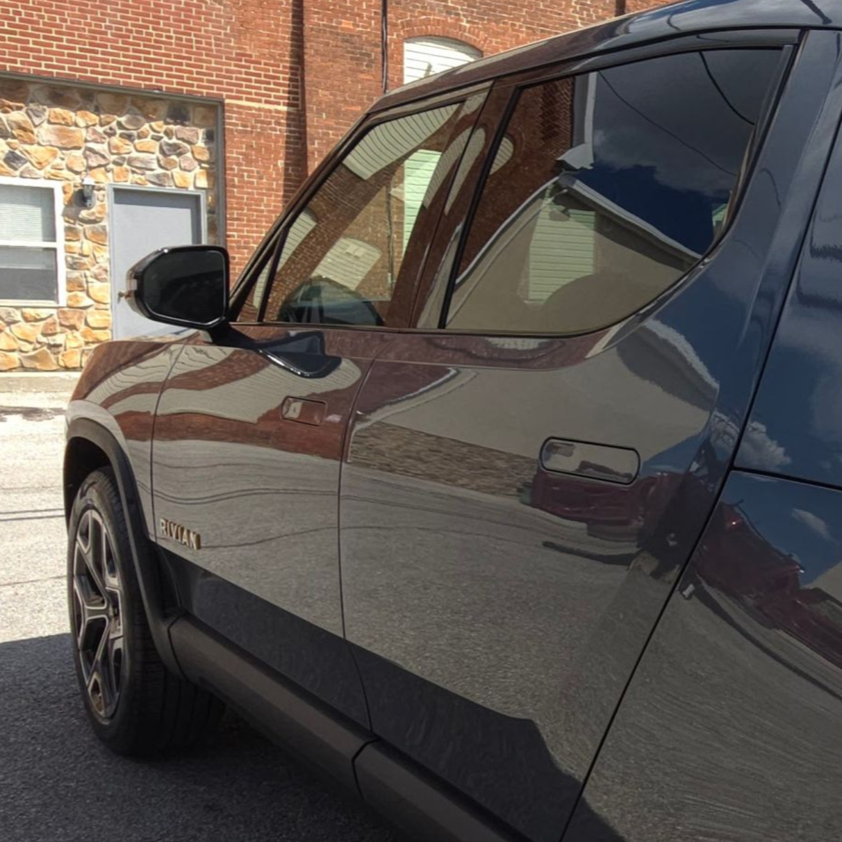 Dark gray Rivian electric vehicle parked on a street, reflecting buildings and sky in its windows.