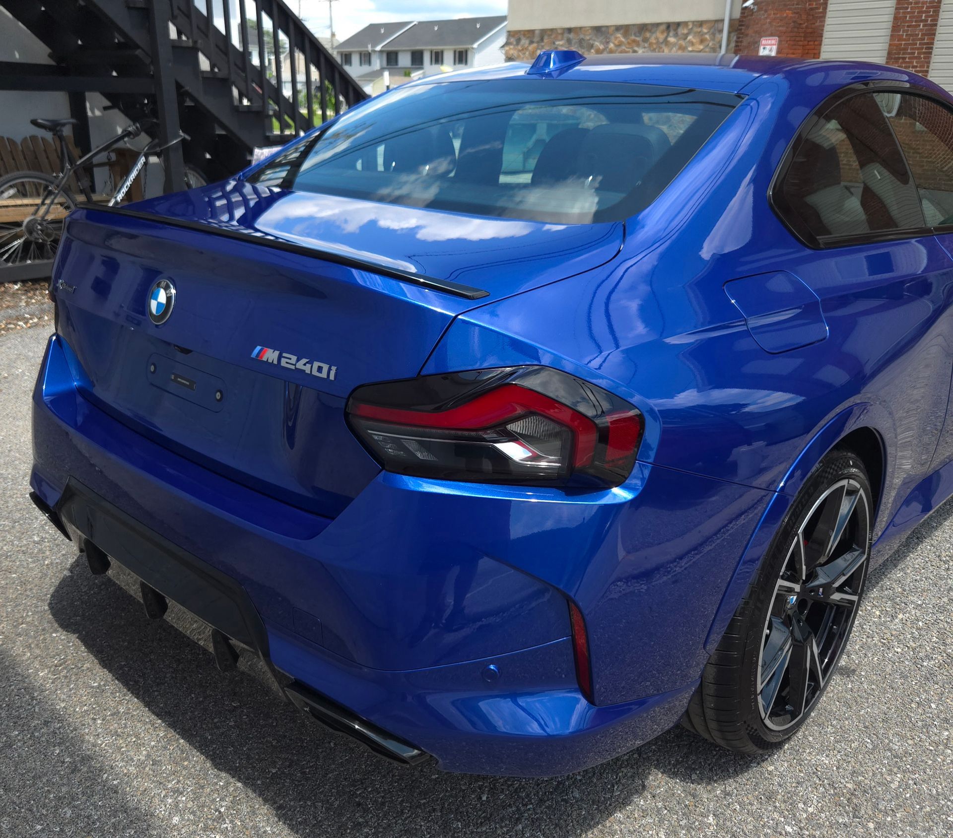 Blue BMW M240i coupe, parked on pavement; black spoiler, taillights, and wheels.