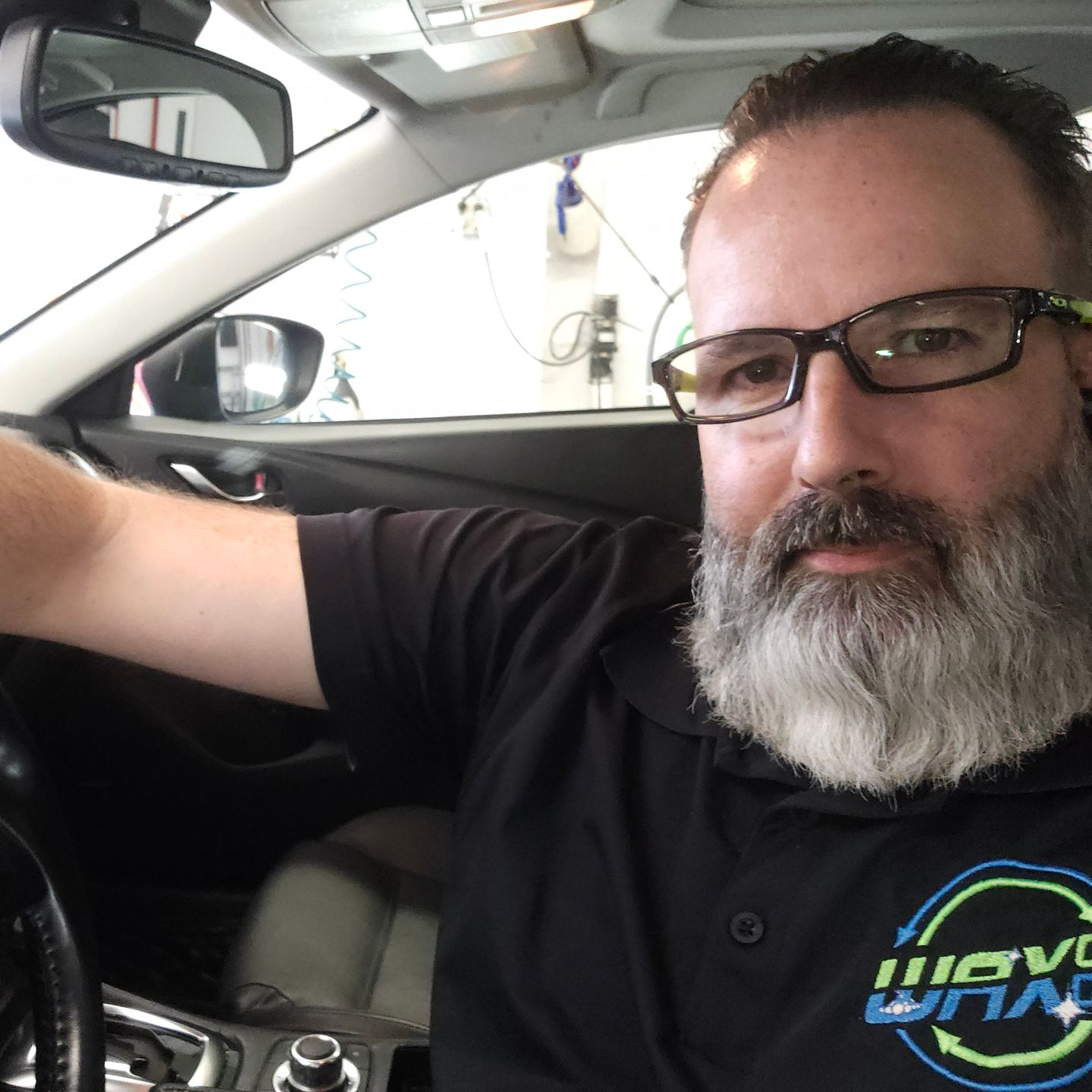 Man with glasses and gray beard in a car, wearing a black shirt with a logo.