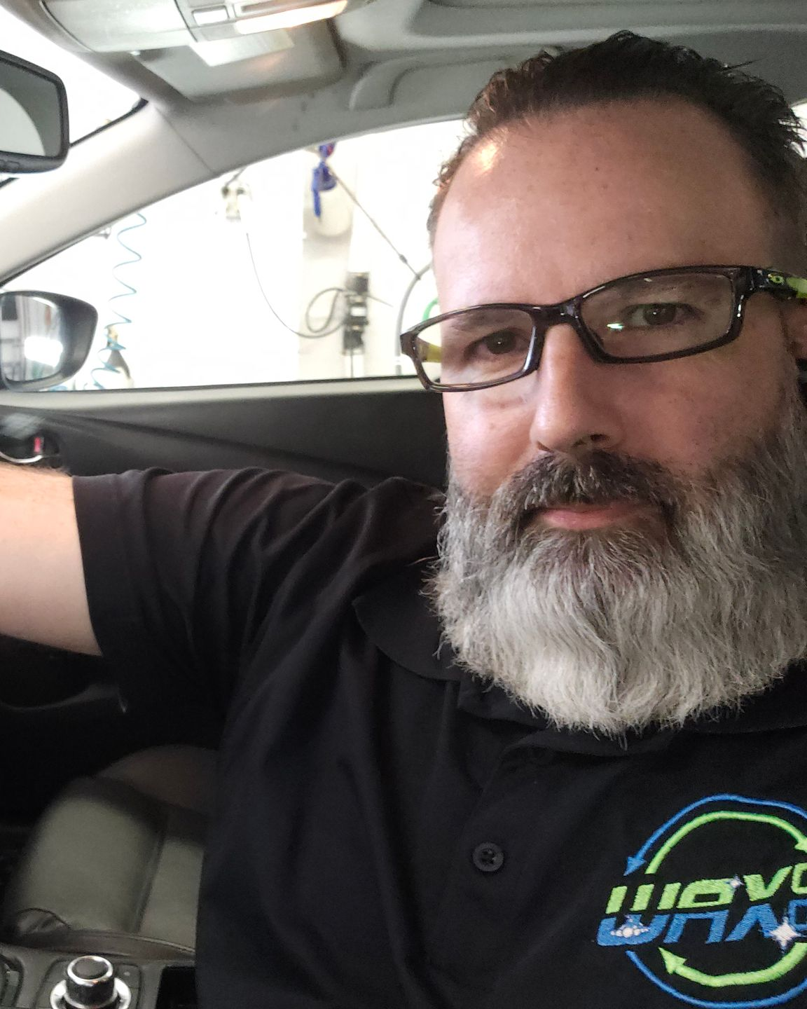 Man with glasses and beard in a car, wearing a black shirt with logo.