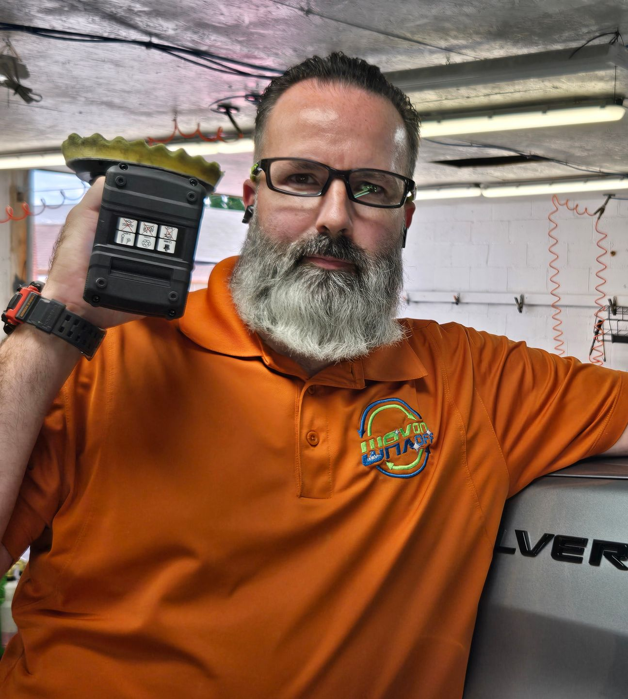 Man with glasses and beard holding a car detailer, in an auto shop. He's wearing an orange shirt.