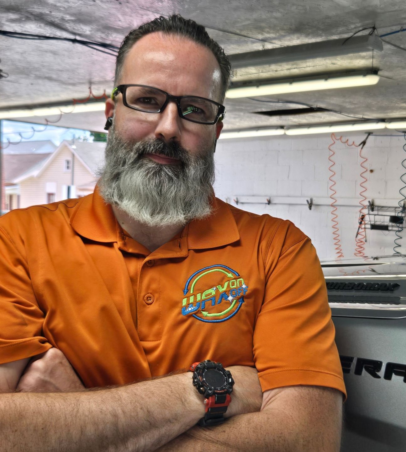 Man with glasses and a beard in an orange shirt, arms crossed. Inside a shop.
