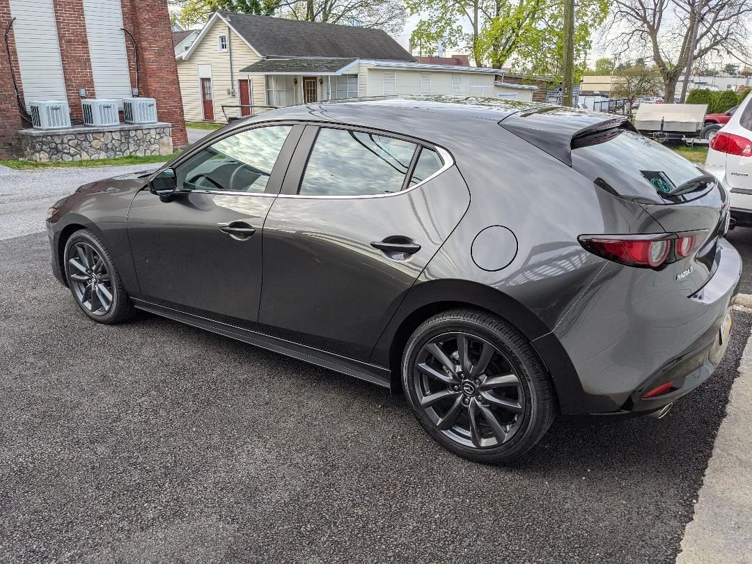 Gray Mazda 3 hatchback parked on gravel in front of a building. Black wheels.