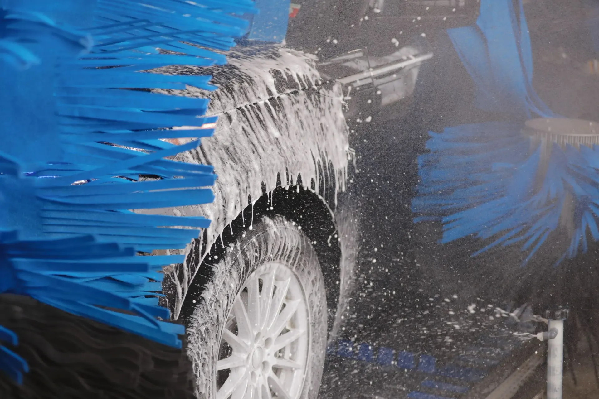 Car being washed at a car wash, covered in soap and water sprays, with blue brushes.