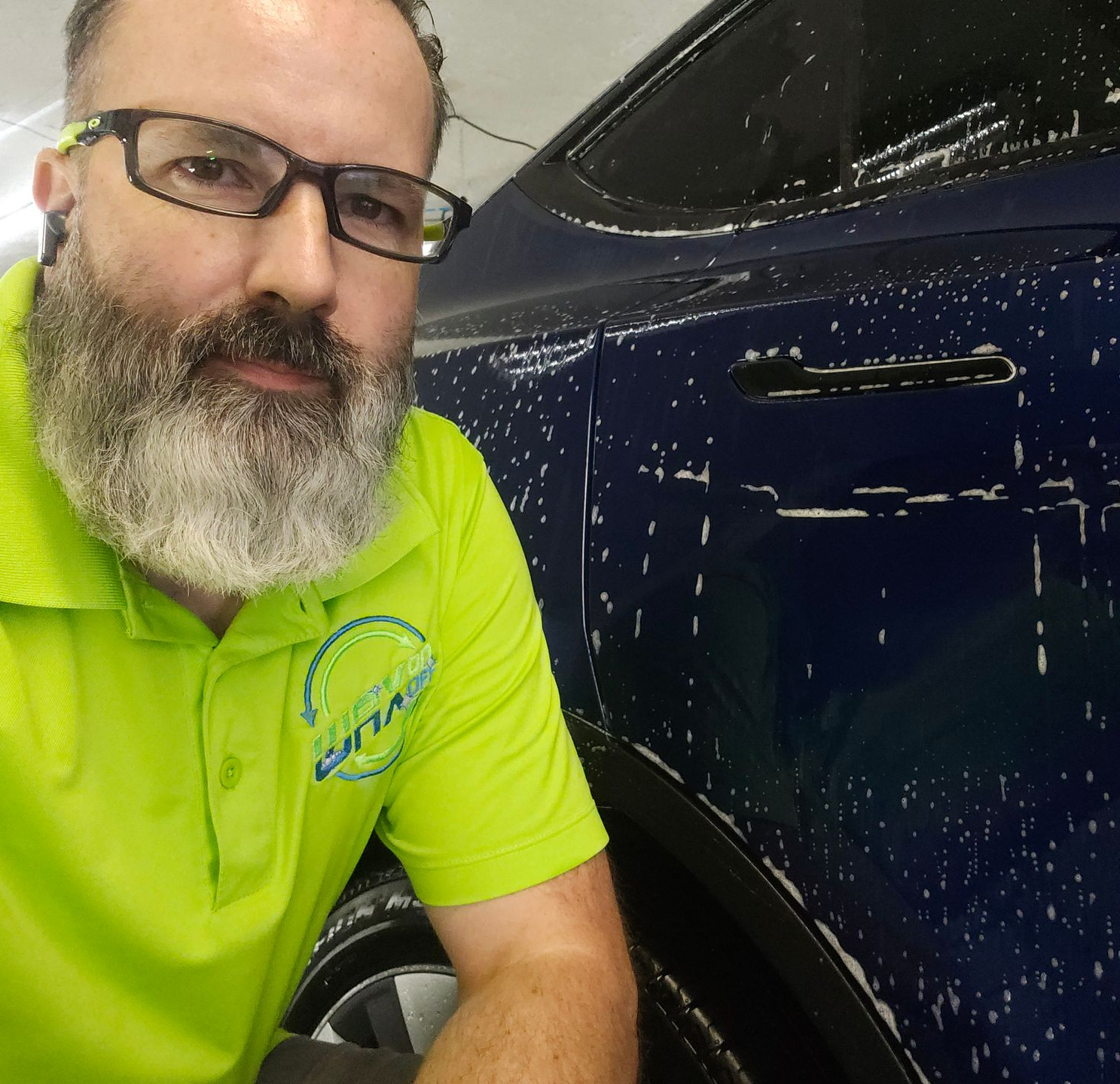 Man with beard wearing glasses and a green shirt, washing a blue car.