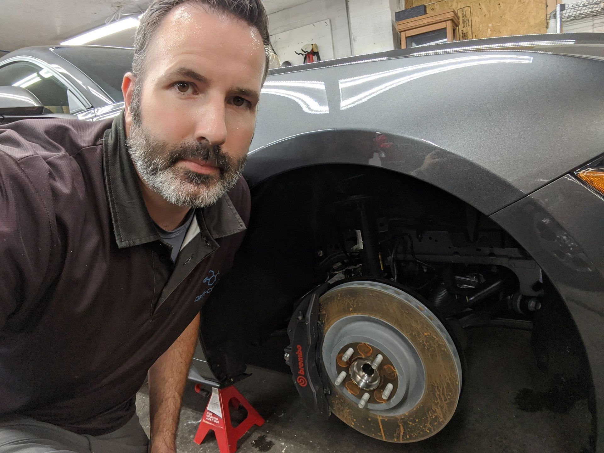 Man with a beard by a car with a removed wheel; in a shop.