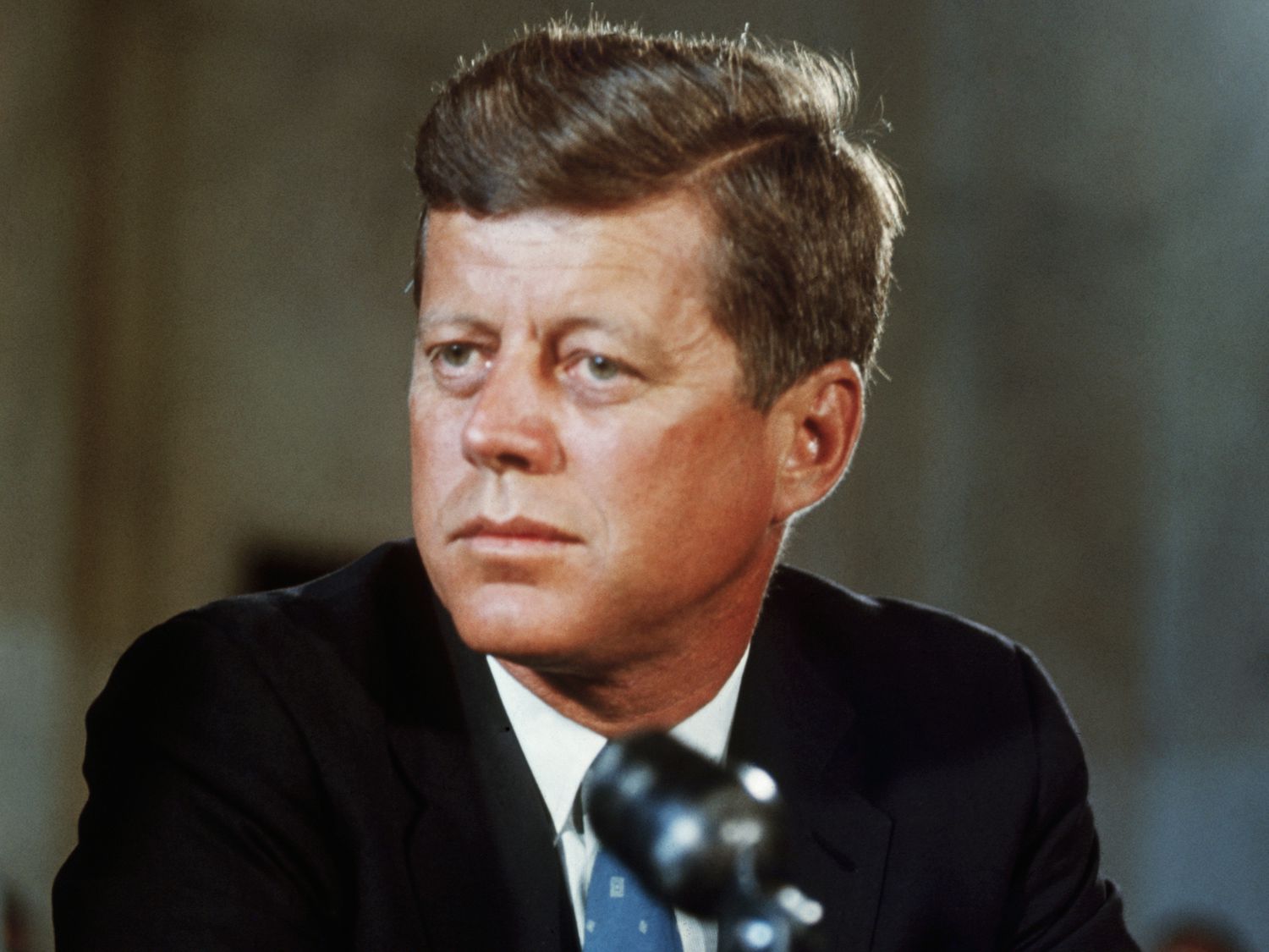 John F. Kennedy in a suit, looking serious at a microphone.