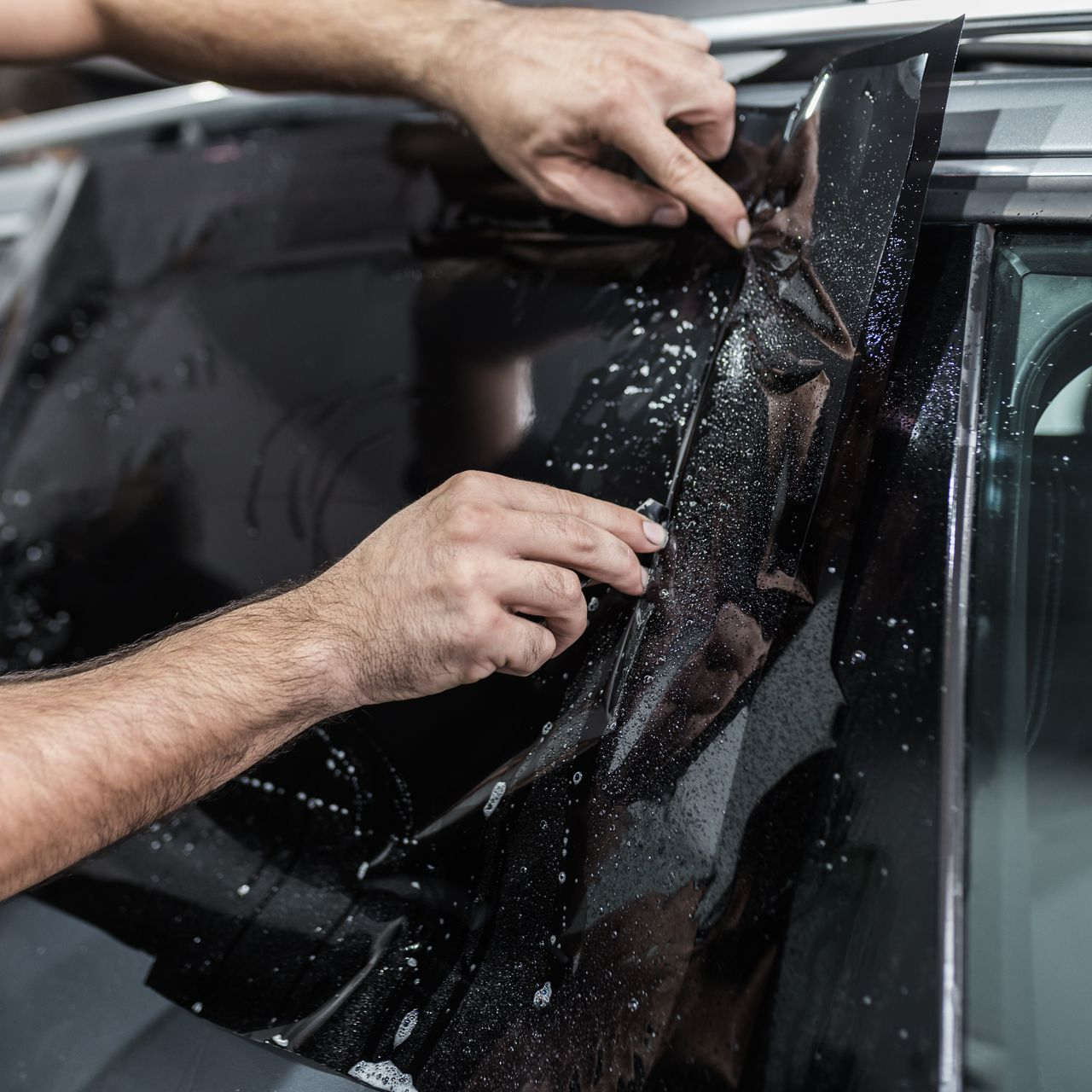 Hands applying tinted film to a car window; workshop setting.
