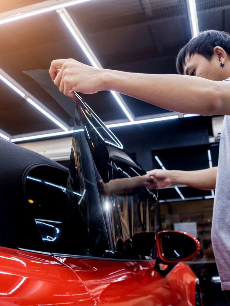 Man applying black tint to the side window of a red car inside a well-lit garage.