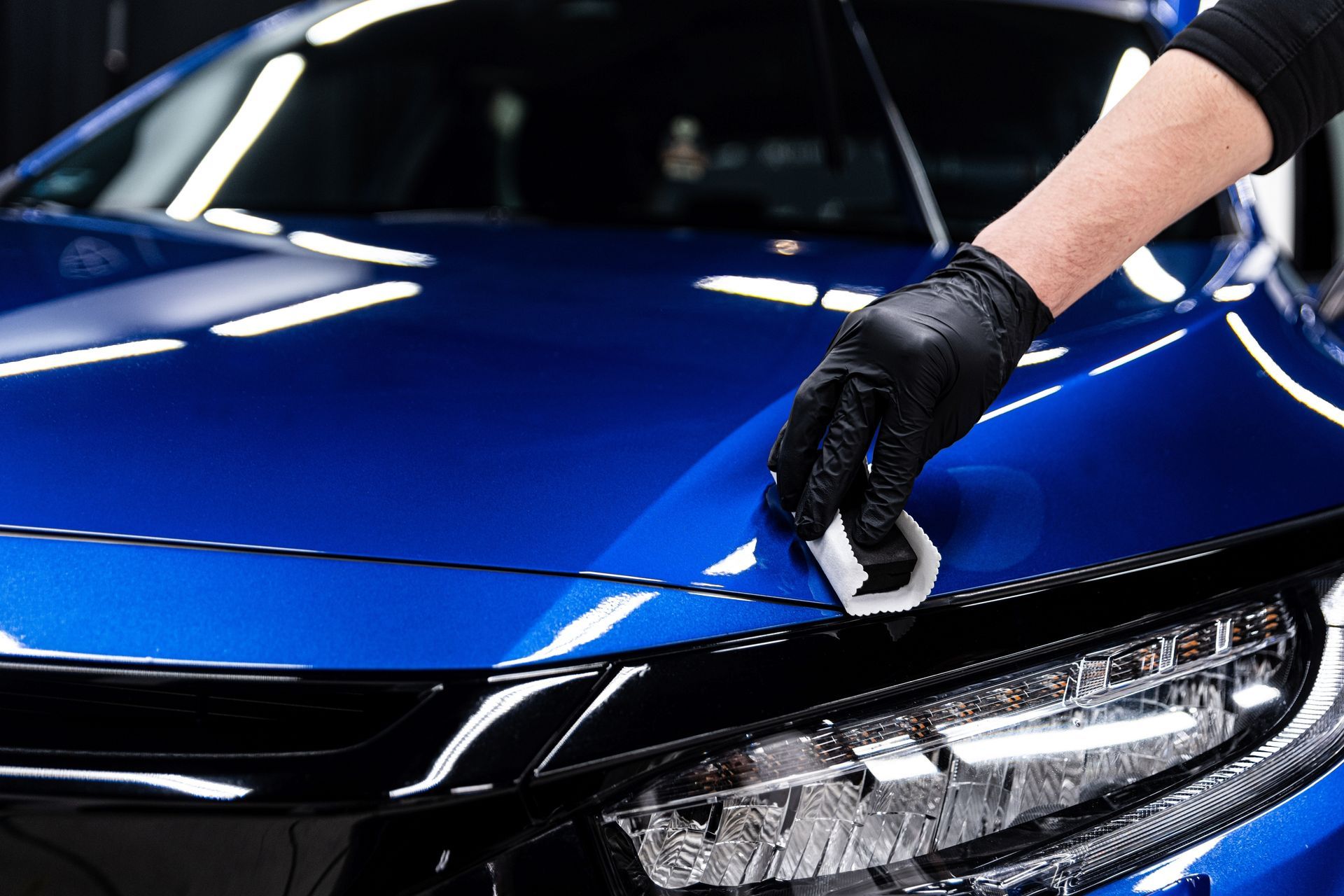 Hand in black glove applying ceramic coating to blue car hood.