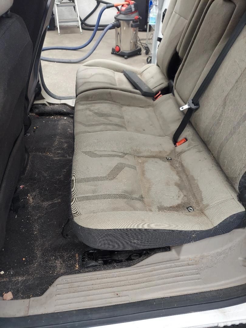 Dirty car back seat being cleaned with a vacuum. Gray seats and black floor with water stains.
