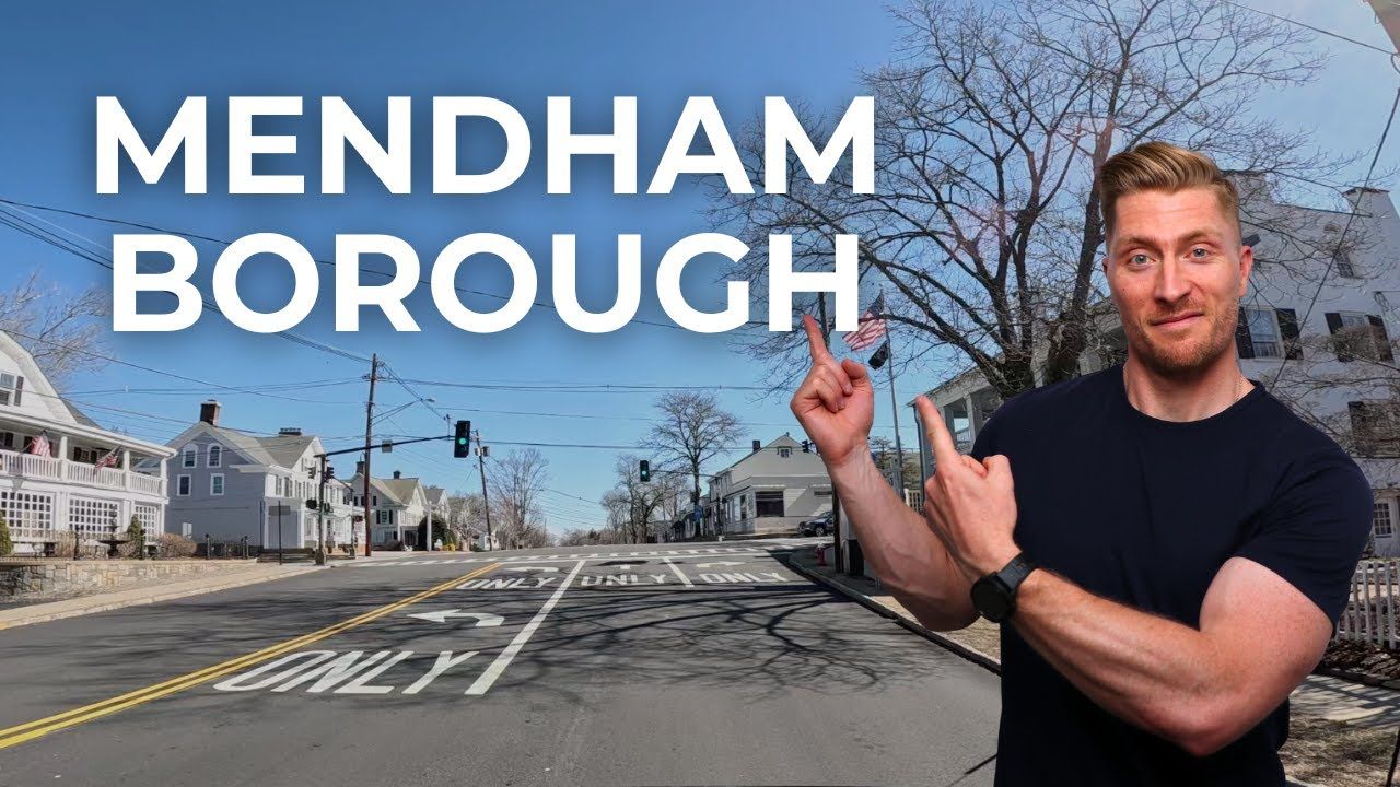 Man pointing towards a street in Mendham Borough, with buildings on either side and blue sky.