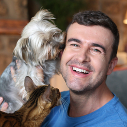 A smiling person in a blue shirt is being nuzzled by a fluffy dog while a brown tabby cat sits near their shoulder.