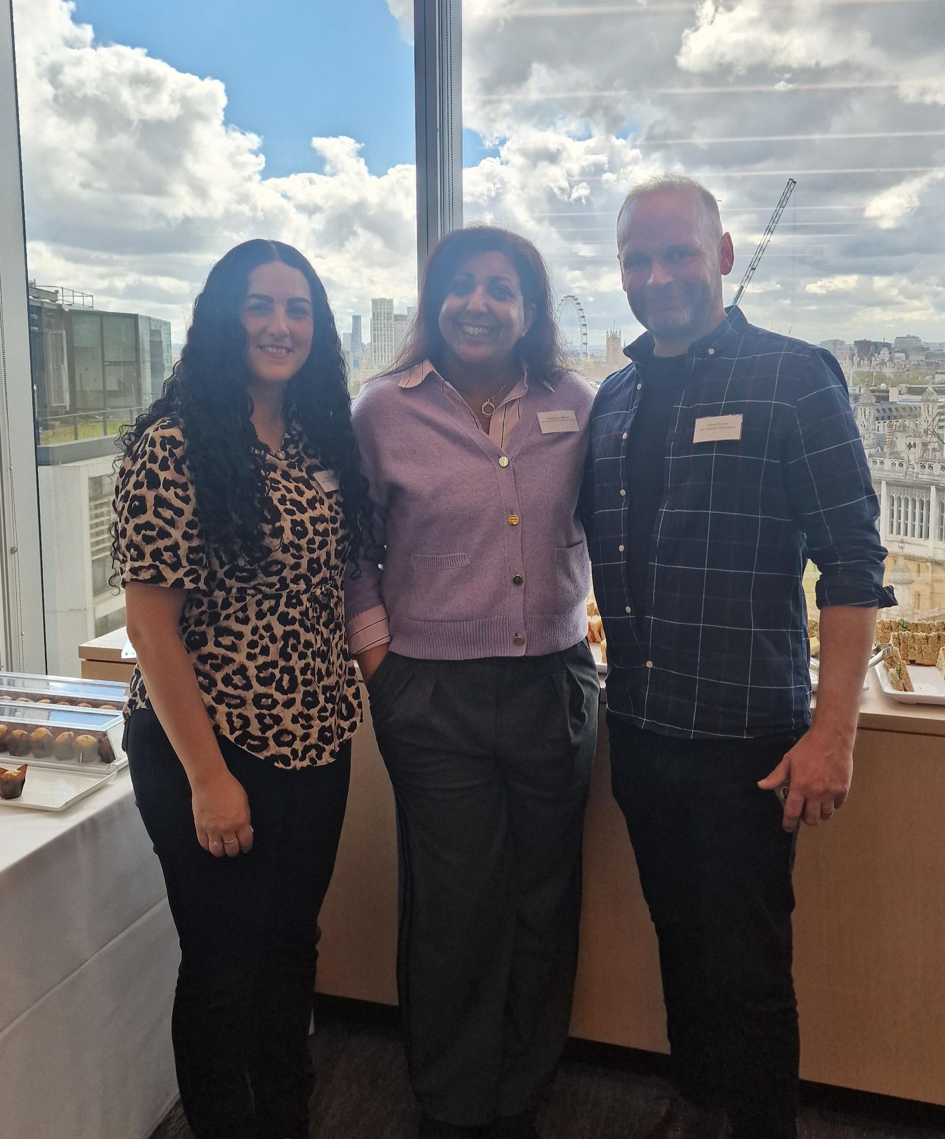 A smiling person in a dark coat holds a small gray puppy in front of a yellow Birmingham Dogs Home sign.