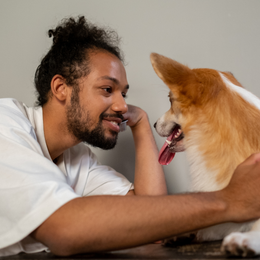 A person with a beard and hair in a bun lies on the floor, smiling affectionately at a Corgi with its tongue out.